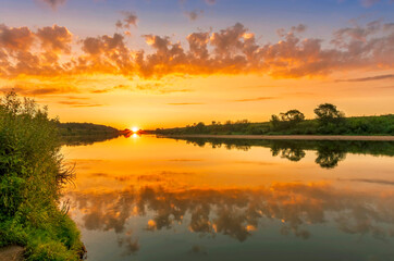 Plakat Scenic view at beautiful summer river sunset with reflection on water with green bushes, calm water ,deep colorful cloudy sky and glow on horizon on a background, spring evening landscape