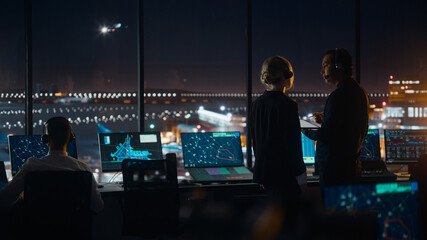 Female and Male Air Traffic Controllers with Headsets Talk in Airport Tower at Night. Office Room Full of Desktop Computer Displays with Navigation Screens, Airplane Flight Radar Data for Controllers. © Gorodenkoff
