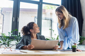 Positive businesswoman looking at african american colleague with paper folder in office