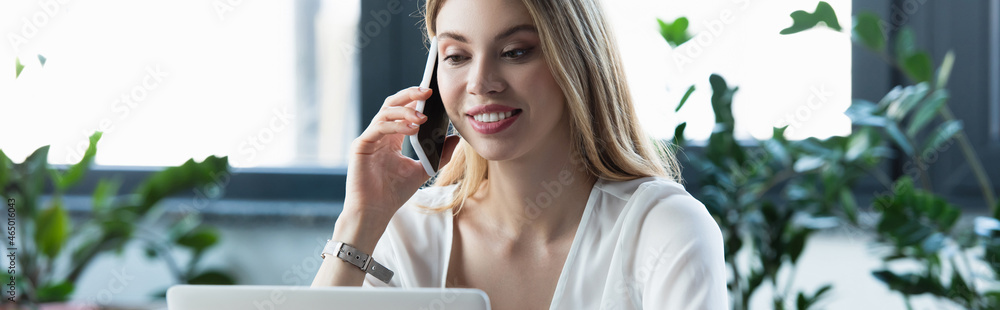 Wall mural happy businesswoman talking on smartphone near laptop in office, banner