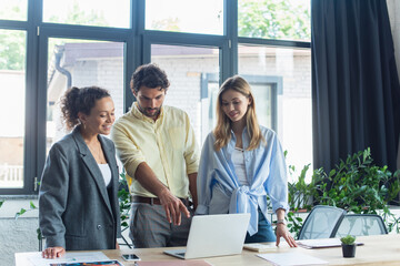 Businessman pointing at laptop near smiling interracial businesswomen in office