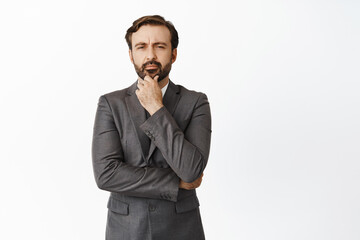 Thoughtful corporate man makes decision, thinking and looking at camera, making choice, standing in grey suit over white background