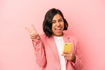 Young latin woman holding mobile phone isolated on pink background joyful and carefree showing a peace symbol with fingers.