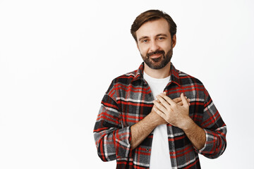 Portrait of smiling bearded man holding hands on heart and looking caring and touched, standing over white background