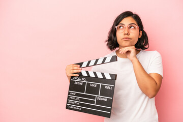 Young latin woman holding clapperboard isolated on pink background looking sideways with doubtful and skeptical expression.