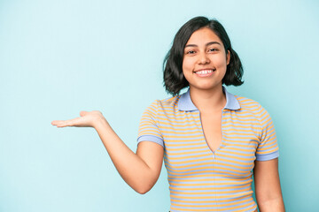 Young latin woman isolated on blue background showing a copy space on a palm and holding another hand on waist.
