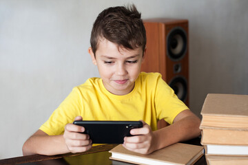 a boy of European appearance is sitting at a table with books and looking at the phone. The concept of modern children