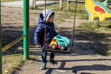 happy joyful kid is playing at playground area with toy plastic truck and swings.swing with massive chains.little boy with hood on head and letters print on clothes.autumn,fall,sunny day