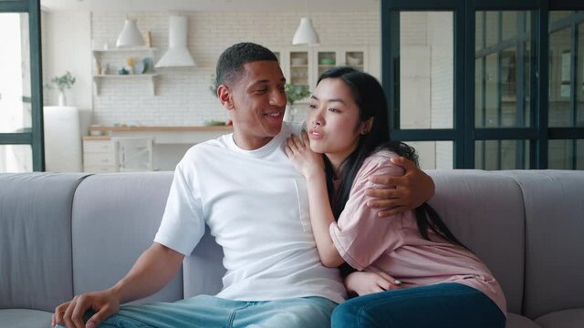 Young asian woman leaning on the shoulder of her african american boyfriend, relaxing on the couch at home on her day off, discussing together, looking away, enjoying spending time together indoors