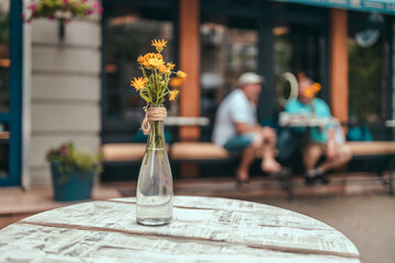 Wooden table with a transparent glass vase with yellow flowers. Street urban cafe