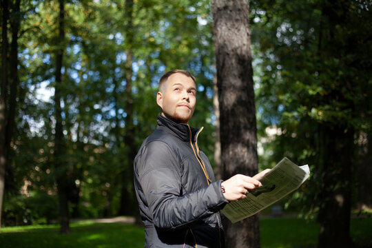 Caucasian young man reading newspaper outdoors in autumn jacket. Guy reads news in green park and looking away with hope for the future. Ecology news concept. Good news about environment.