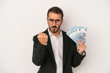 Young caucasian business man holding banknotes isolated on white background showing fist to camera, aggressive facial expression.