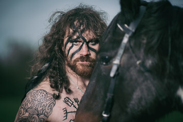 Obraz premium Medieval warrior berserk Viking portrait with tattoo on skin, cuts on body and braids in hair with weapons to attacks enemy. Concept historical photo