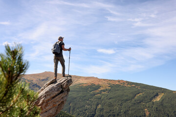 Man with backpack and trekking poles on rocky peak in mountains, back view