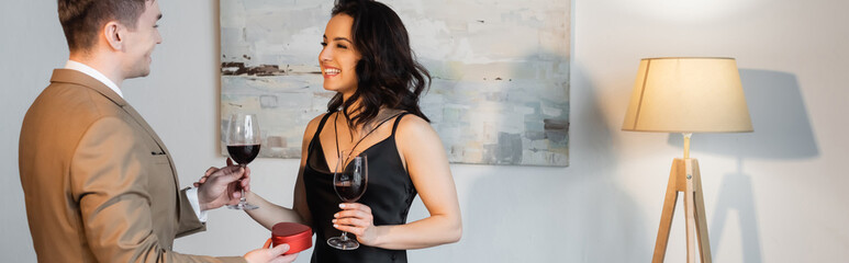 happy man holding heart-shaped gift box and glass of wine near smiling girlfriend in black slip dress, banner