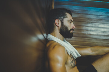 Obraz na płótnie Canvas Beautiful healthy boy relaxing in a woden sauna. Young bearded man resting after workout in the gym. Fitness male with tatoo and white towel, laying on the bench in the spa. Wellness, health concept