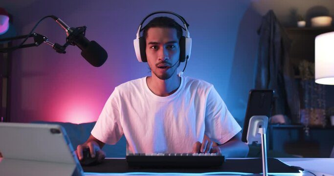 Young Asian man playing online computer video game, colorful lighting broadcast streaming live at home. Ecstatic celebration winning a match. Gamer lifestyle, E-Sport online gaming technology concept