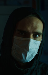 The face of a man wearing a medical mask and a black hood. Dark background.