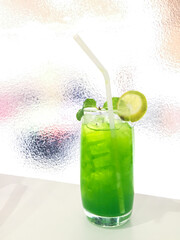 Lime green soda with ice in classic glass with white straw on white table and glass background