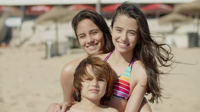 Happy sisters and brother sitting together on sandy beach. Front view of smiling kids in swimsuits looking at camera, having good time together during summer vacation at sea. Family,emotion concept