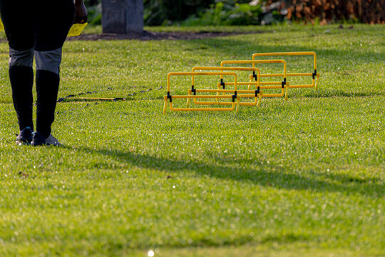 Sport and recreation concept, Selective focus yellow football obstacle training hurdle in a row with blurred trainer on green grass filed, Outdoor sport practice on meadow with sunlight in the morning