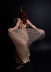 Full length  portrait of red haired  girl wearing a creamy fantasy gown like a fairy goddess costume.  standing  pose with elegant gestural movement  backwards the the camera, isolated on dark studio 