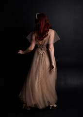 Full length  portrait of red haired  girl wearing a creamy fantasy gown like a fairy goddess costume.  standing  pose with elegant gestural movement  backwards the the camera, isolated on dark studio 