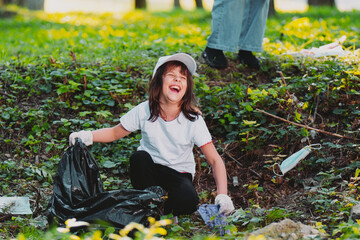 Close up photo of a laughing young girl sitting down on the ground in white gloves holding a garbage bag and picking up plastic rubbish.