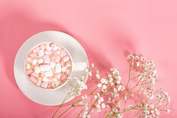 Cup of cocoa with marshmallows and white flowers on a pink background, top view, free space for text