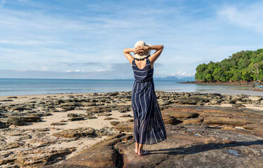 Asian girl in a dress and a hat stands on the beach looking at the sea enjoying her vacation. 