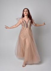 Fototapeta premium Full length portrait of red haired girl wearing a creamy fantasy gown and crystal crown, like a fairy goddess costume. standing pose with elegant gestural arm movement , isolated on light studio b