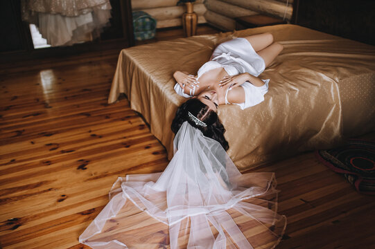 A beautiful, sexy, stunning bride in white lingerie, underwear with a long veil lies on the bed in the room, the interior of the hotel in a wooden house and poses. Wedding photography.