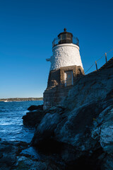 Fototapeta na wymiar White lighthouse on the rocky cliff over the blue ocean water against clear sky. Castle Hill Lighthouse in Newport, Rhode Island.