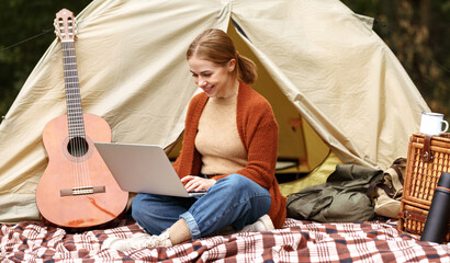 Young smiling woman sitting near tent with laptop computer and working online during camping © JenkoAtaman