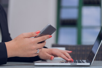 Close-up of a woman's hands, holding a smartphone, typing on her laptop.