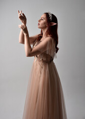 Fototapeta premium Full length portrait of red haired girl wearing a creamy fantasy gown and crystal crown, like a fairy goddess costume. standing pose with elegant gestural hands, isolated on studio background