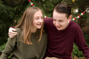 Lifestyle outdoor holiday portrait of young happy brother and sister sitting and laughing with trees and Christmas garland in background