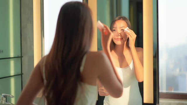 Beautiful young Asian woman looking in mirror of hotel bathroom and cleaning face with morning sunlight. Femininity concept