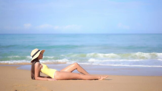 Sive view and woman yellow swimsuit and sunhat resting lying leaning on elbows at a sandy beach near the sea when foamy waves roll over the sand. Beautiful female model on travel vacation holidays
