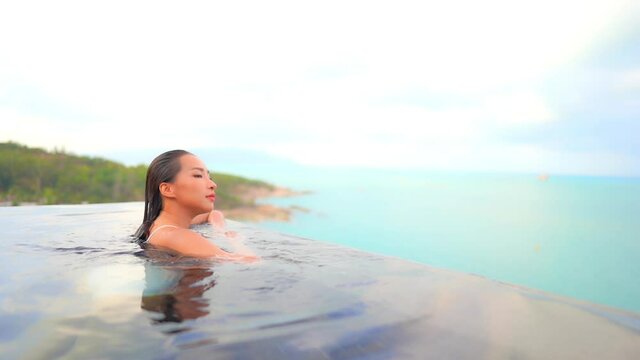 Asian woman swims toward edge of infinity pool overlooking ocean beauty relaxing poolside in fashion swimsuit. Asian wellness