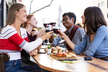 Multiracial friends toasting red wine at the outdoor pub - Food and beverage lifestyle concept with happy people having fun together at pub - Bright filter with focus on the guys in the background