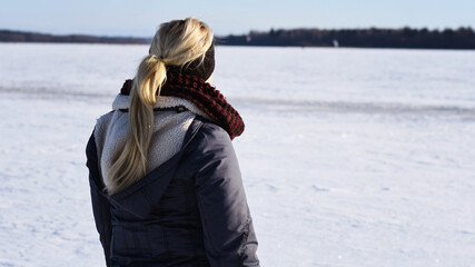 Fototapeta na wymiar Woman with blonde hair in a ponytail wearing warm winter clothes as she stands outside looking out over the winter landscape. Photo taken in Sweden.
