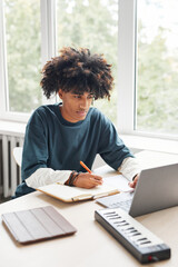 Vertical portrait of African-American teenage boy using laptop while studying by window at home or in college