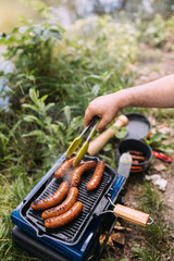 A man on a camping trip preparing food in nature. There are sausages on a grill.