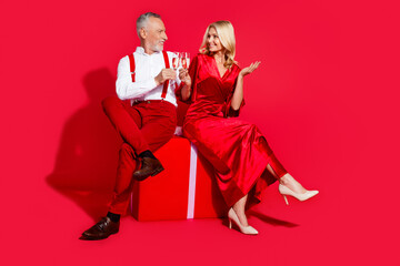 Full length photo of mature couple sitting gift box celebrate eve time noel drink champagne isolated over red color background