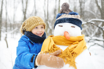 Little boy building snowman in snowy park. Child embracing snowman wearing hat and scarf. Active...