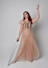 Full length  portrait of red haired  girl wearing a creamy fantasy gown and crystal crown, like a fairy goddess costume.  Standing pose holding a sword weapon  isolated on light studio background.