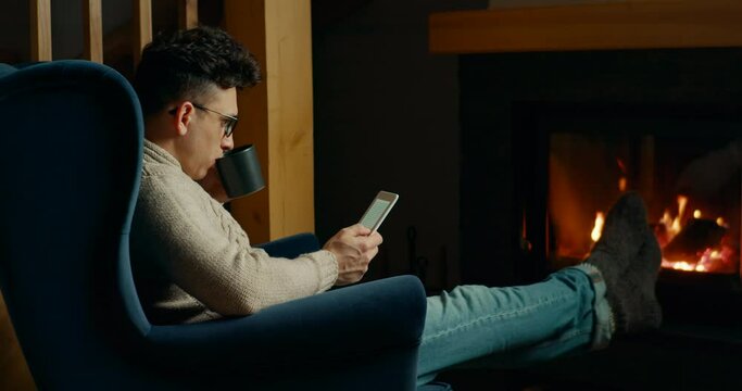 Man Reads eBook on electronic Reader or uses Tablet relaxing at Fireplace in Cozy Room at Winter Night. Christmas Seasonal Leisure or Comfortable Warm Autumn Evening Rest. 4K slider pan wide shot