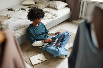High angle portrait of teenage African-American student sitting on floor at home or in college dorm...