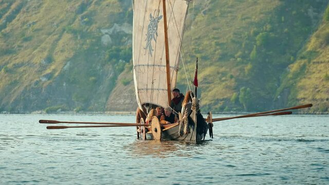 Vikings Sail on an Old Ship with a Raised Sail on a Calm River Against the Backdrop of a Rocky Coast. The Men Row the Oars Diligently Towards Adventure. Medieval Reconstruction.